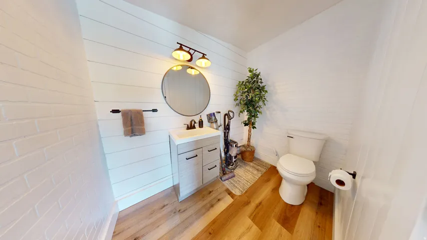 Bathroom featuring wood-finish flooring, a white shiplap accent wall, and a circular mirror above a vanity with integrated sink