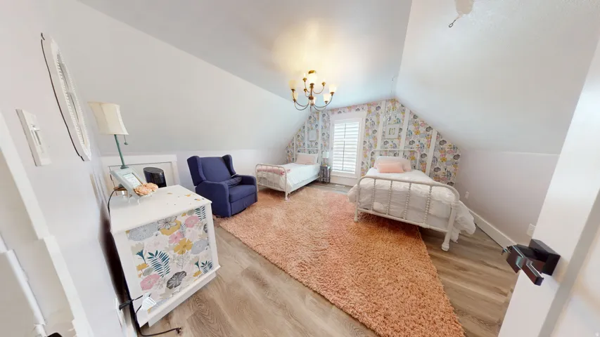Bedroom featuring sloped ceilings, wood-finish flooring, a wall with patterned wallpaper, and a window with white louvered shutters