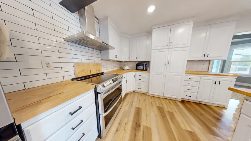 Modern kitchen featuring a stainless steel range with overhead ventilation, light wood-finish countertops, white subway tile backsplash, white shaker-style cabinetry with dark hardware, and wood-finish flooring