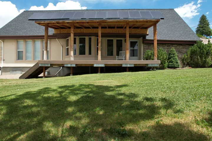 Rear exterior with a covered deck, solar panels, stucco siding, stone veneer accents, and a sloped lawn