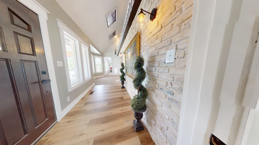Entryway featuring a dark wood front door, wood-finish flooring, a vaulted ceiling, and an exposed brick accent wall with wall-mounted lighting