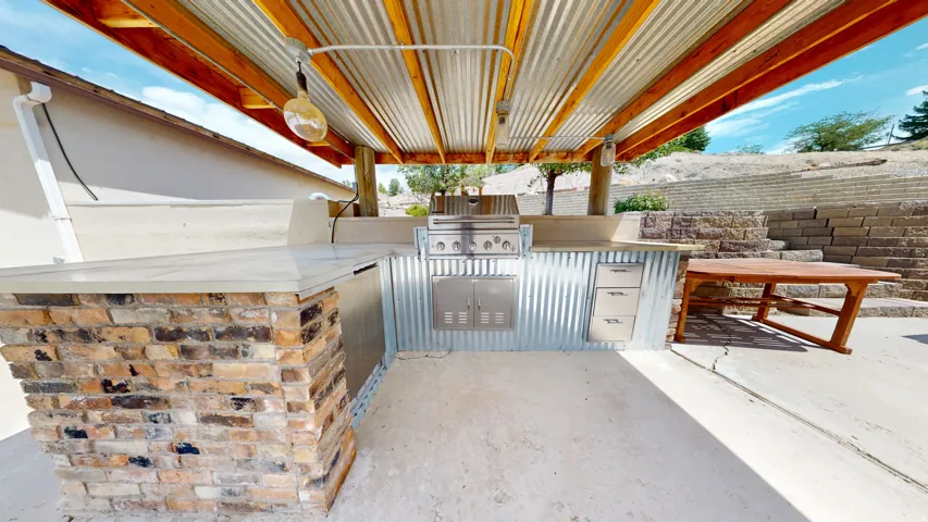 Covered outdoor kitchen featuring a built-in grill, integrated drawers, a brick-clad counter base, and corrugated metal accents