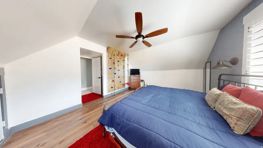 Angled ceiling room featuring wood-finish flooring, a ceiling fan with wood-toned blades, a wall-mounted climbing feature, and a window with white plantation shutters