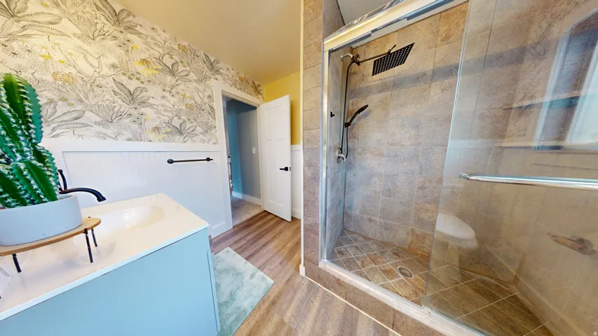 Bathroom featuring a glass-enclosed shower with rain showerhead, tiled shower walls, wood-finish flooring, a vanity with an integrated sink, and decorative wall paneling with botanical wallpaper