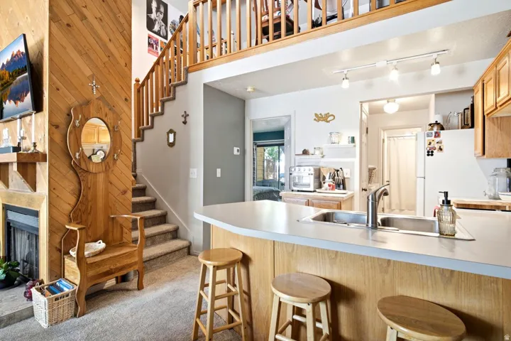 Open-concept living area featuring a kitchen island with integrated sink, wood-finish cabinetry, a fireplace with a wood mantel, a carpeted staircase with a wood railing, and a loft space