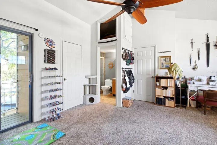 Carpeted interior space featuring a sliding glass door to a private balcony, a ceiling fan with wood-finish blades, and a doorway to a bathroom with a visible toilet and vanity
