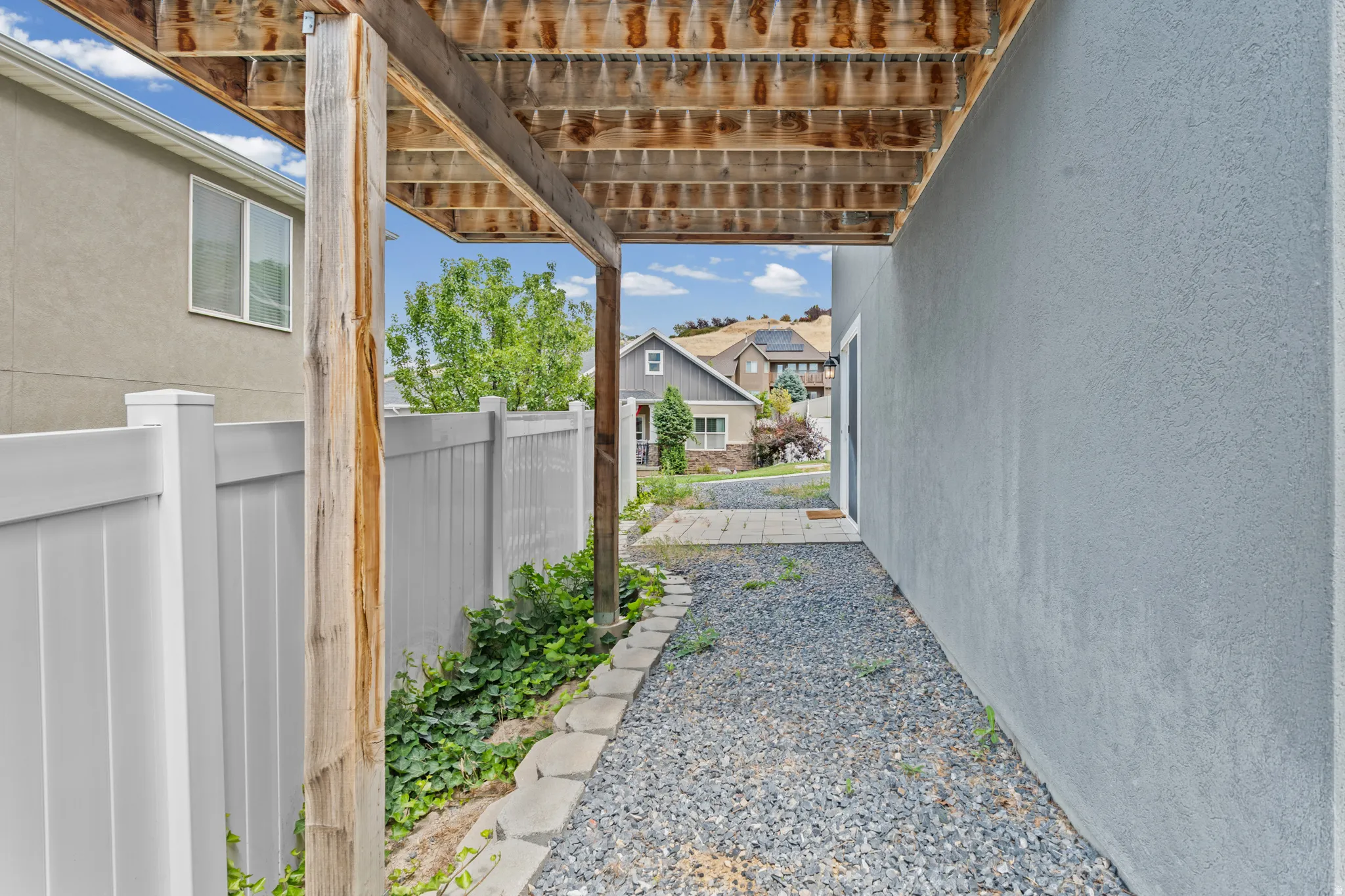 Gravel pathway under a wood-framed deck, featuring a stucco exterior wall and white privacy fencing