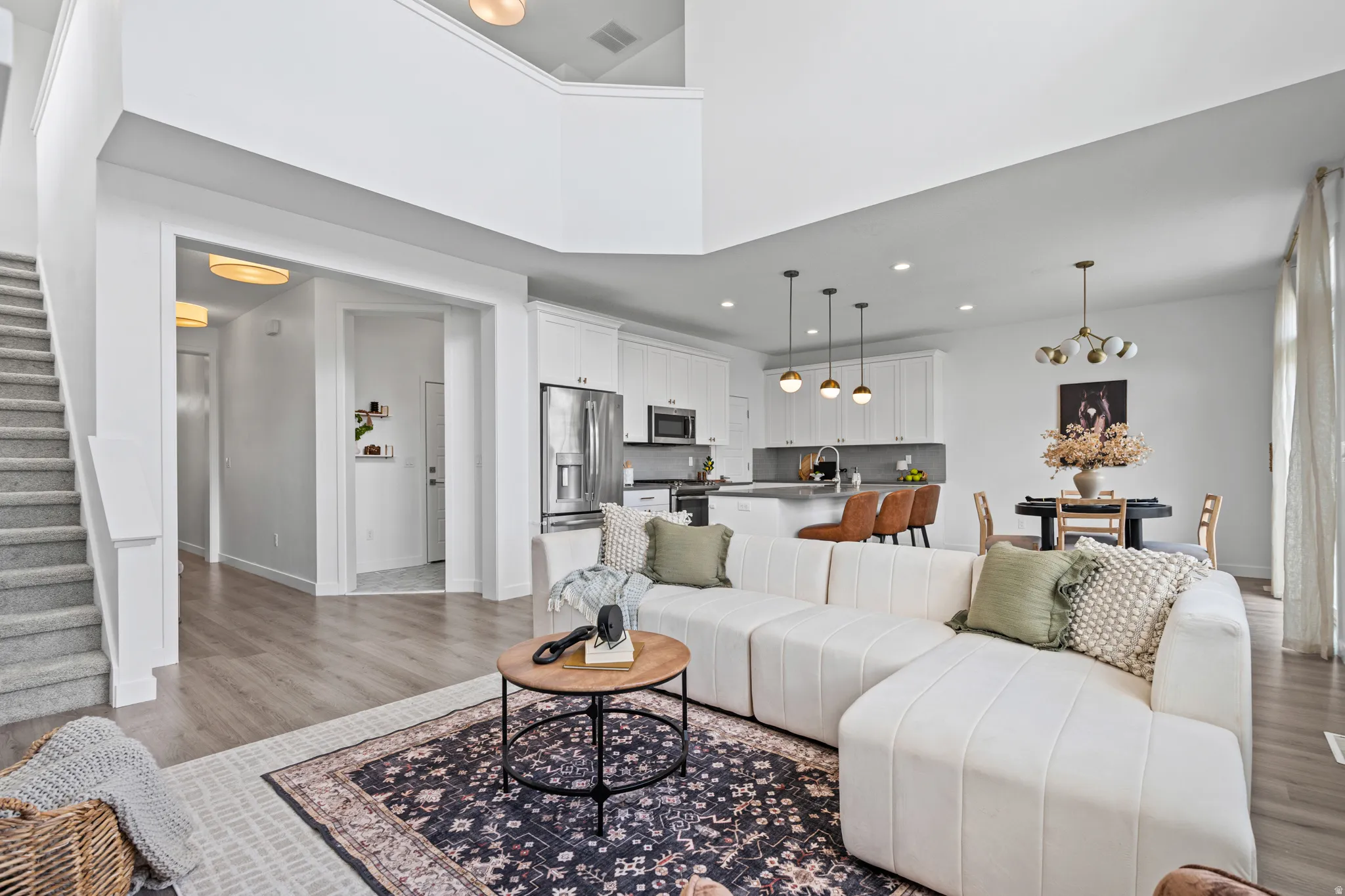 Open-concept living space featuring wood-finish flooring, a two-story ceiling, a carpeted staircase, a kitchen with white cabinetry and stainless steel appliances, and a dining area with a chandelier