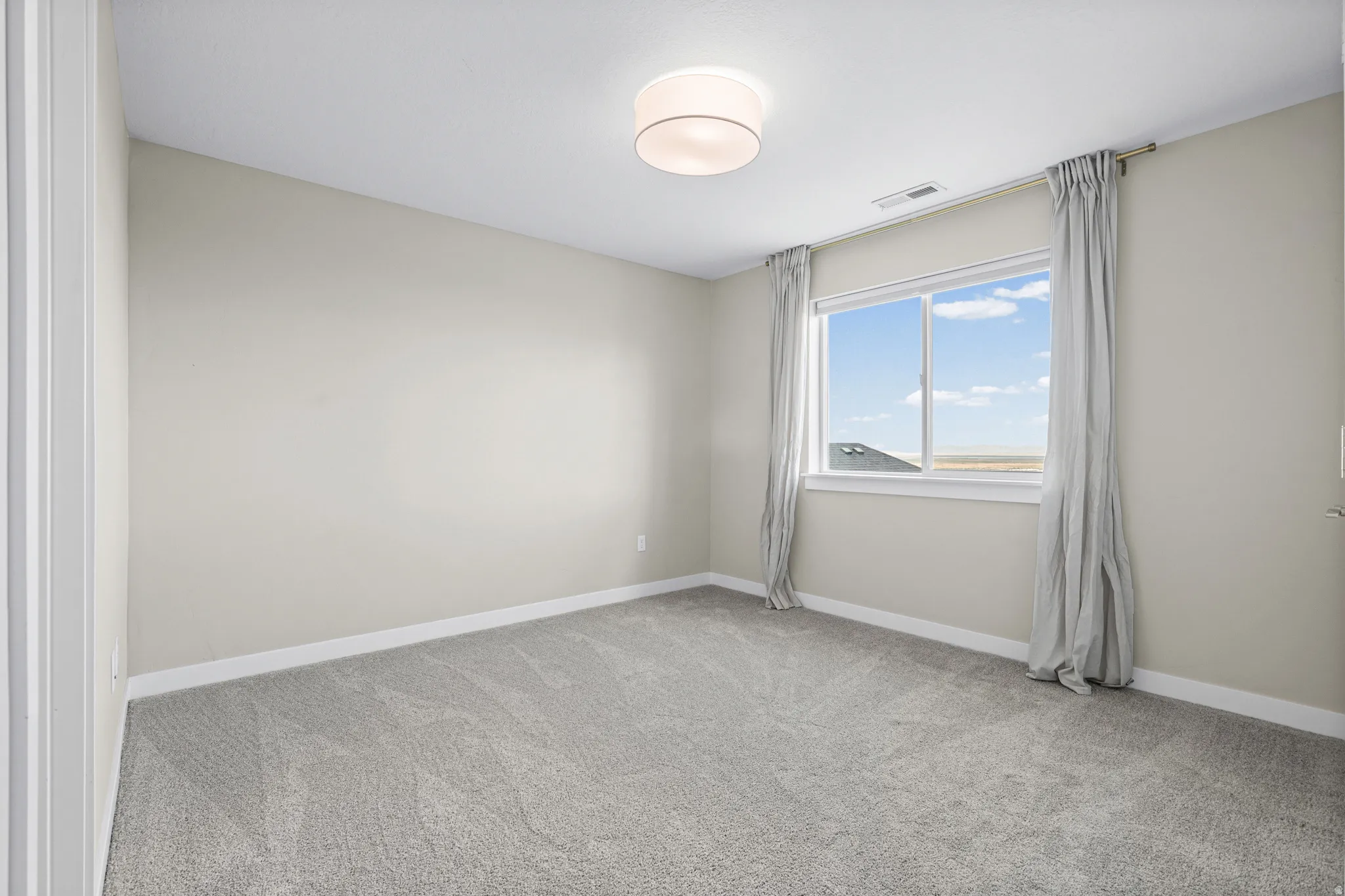 Neutral-toned room featuring a large window with distant scenery, light gray wall-to-wall carpeting, white baseboards, and a contemporary ceiling light fixture