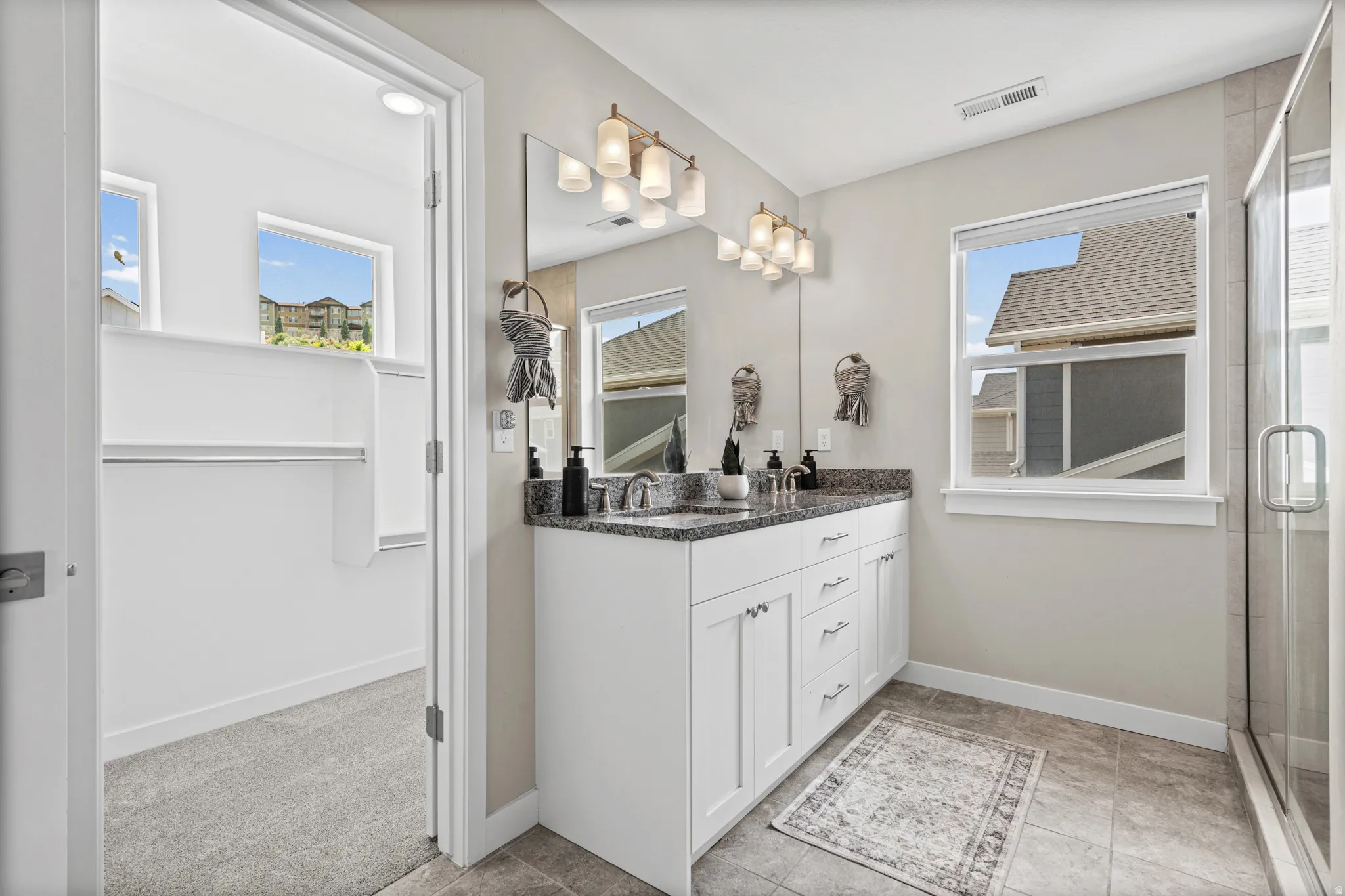Bathroom featuring a dual vanity with a granite countertop, white cabinetry, and a mirrored wall with a multi-bulb light fixture