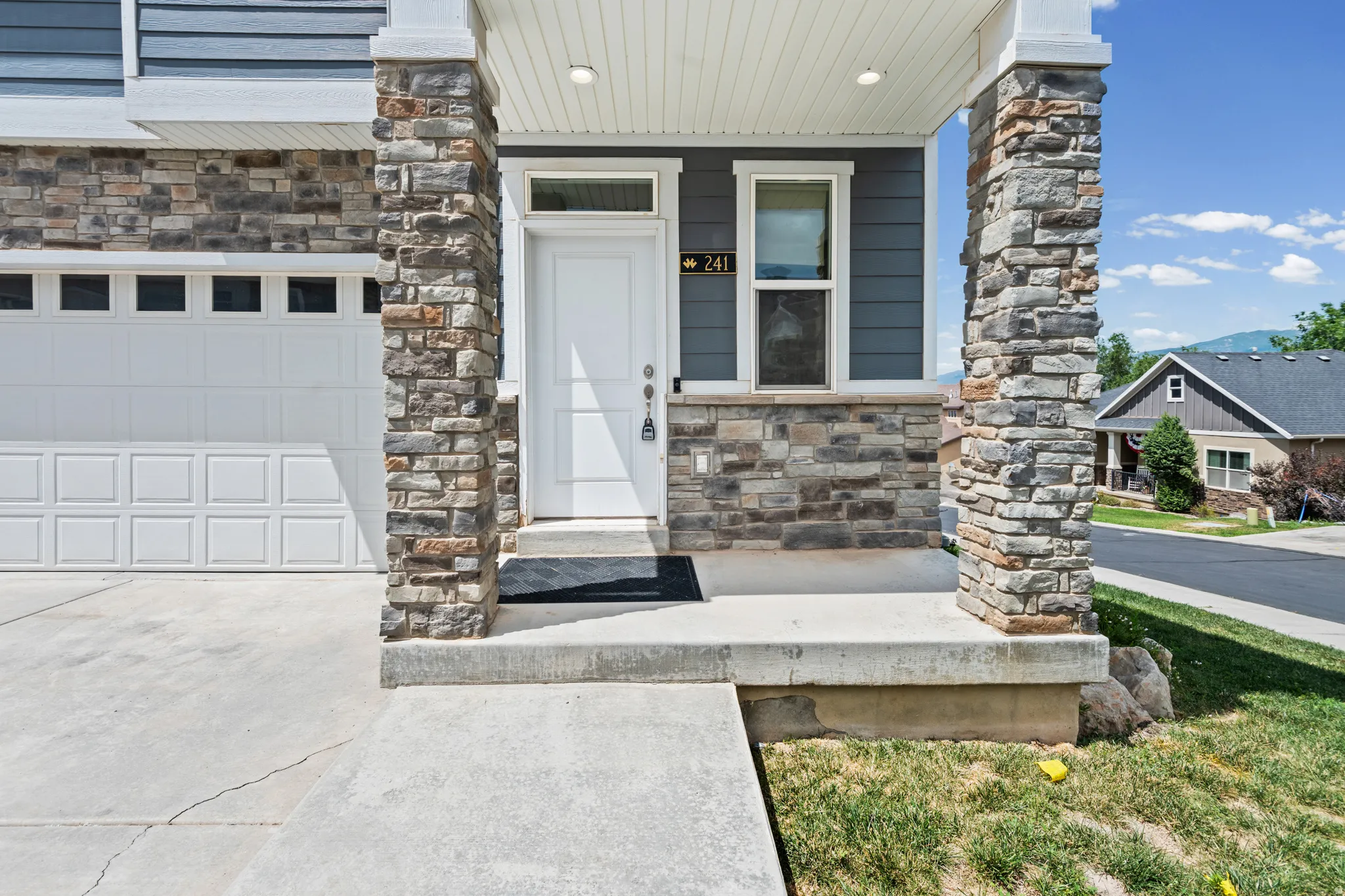 Exterior featuring a white front door with transom window, stone veneer accents, dark siding, white garage door with upper window panels, and a concrete driveway