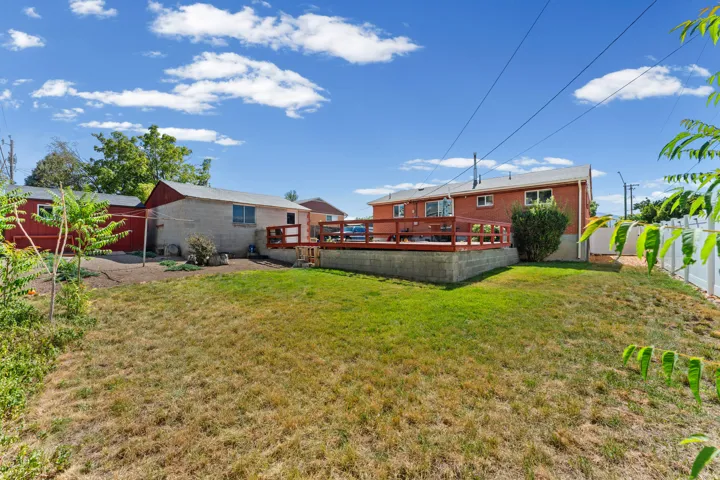 Expansive rear yard featuring a large wood deck with railing, a red brick exterior, multiple windows, a white vinyl fence, and a separate structure with a red barn-style roof