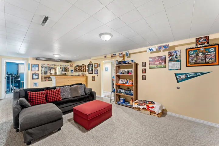 Finished basement living area featuring neutral carpeting, a drop ceiling with recessed lighting, and a built-in wood-finish bar with mirrored backsplash