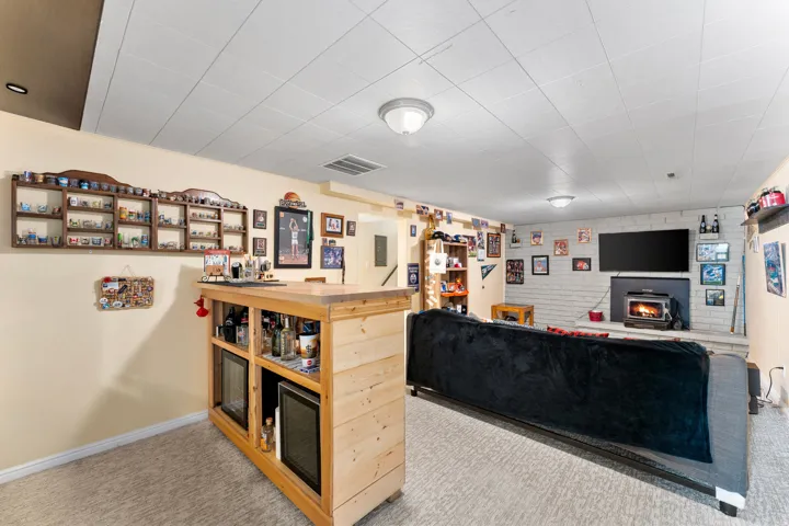 Finished lower level featuring a built-in bar with integrated under-counter refrigerators, a wood-burning stove set against a painted brick fireplace, recessed and flush-mount lighting, and carpet flooring