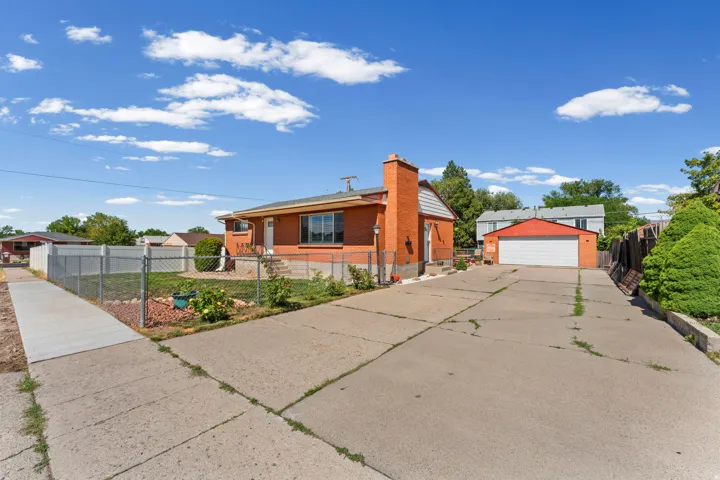 Brick exterior property featuring a paved driveway, front yard with chain link fencing, mature landscaping, and a detached two-car garage with an orange roof