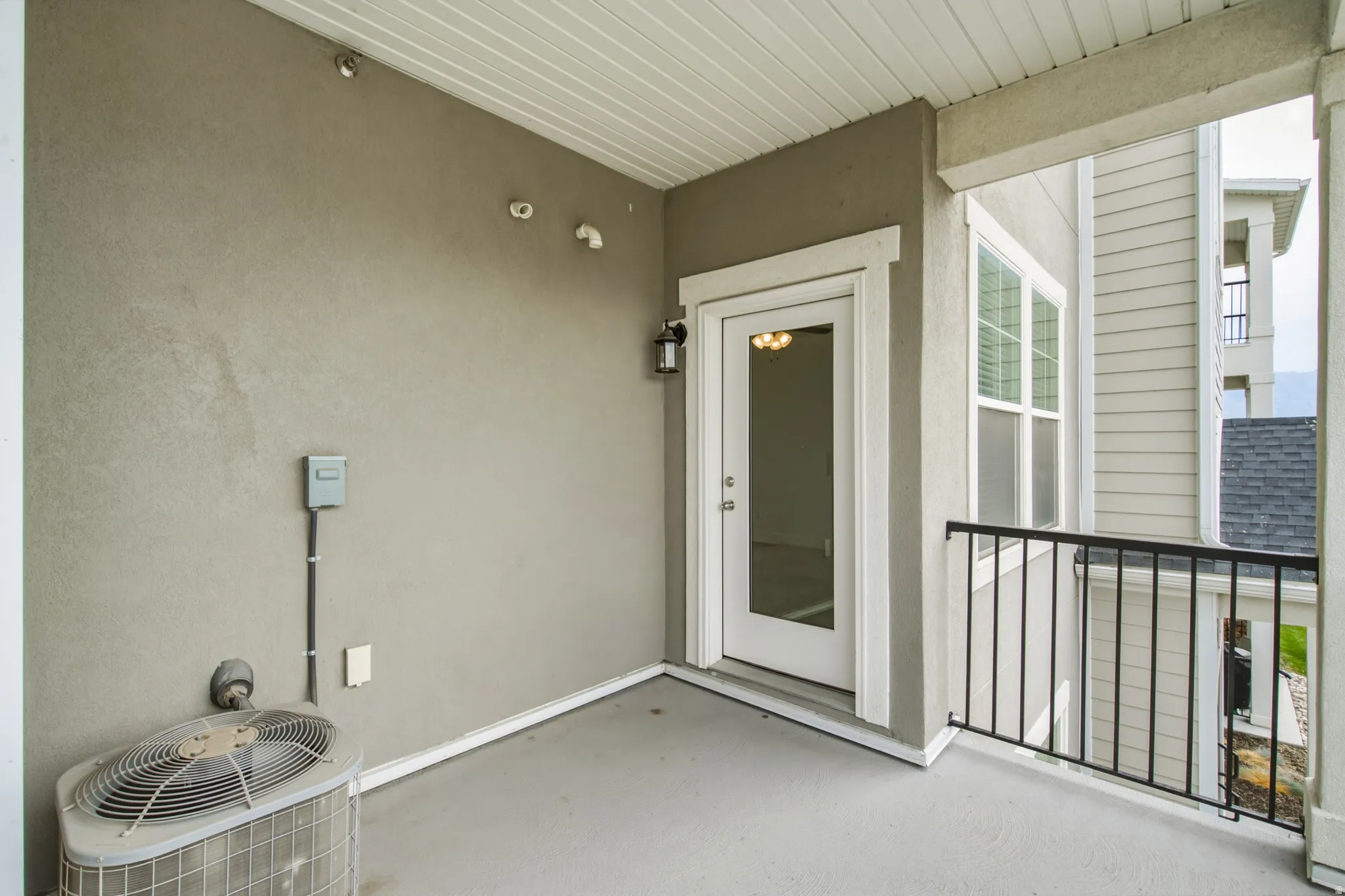 Covered outdoor patio with a stucco wall finish, a white paneled door with a glass insert, a black metal railing, and an overhead lighting fixture