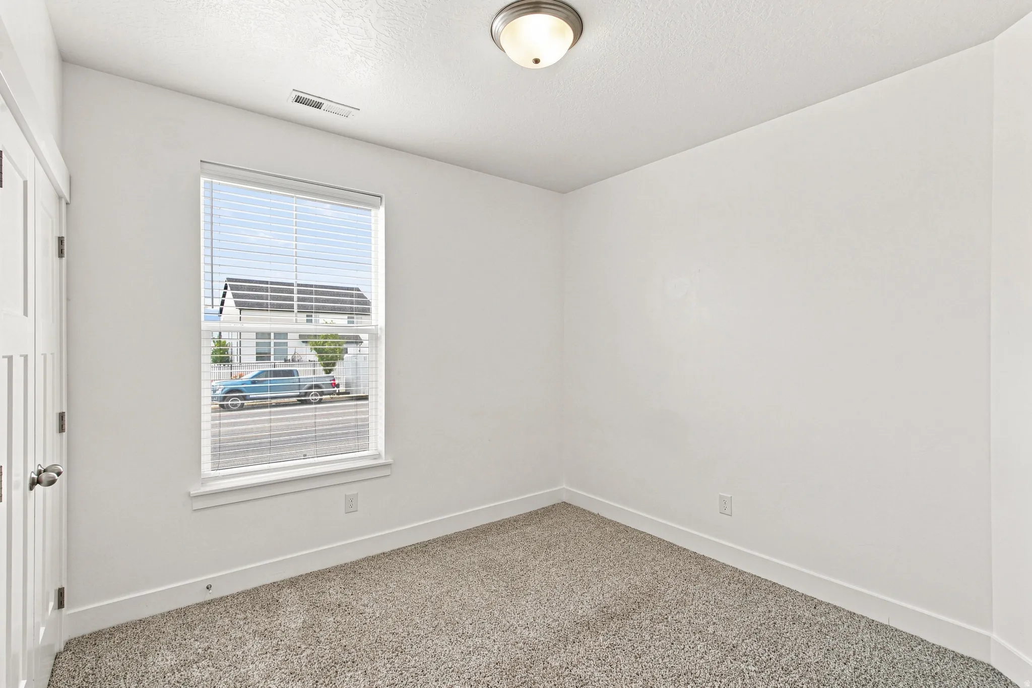 Light-filled room featuring a single window with blinds, neutral wall paint, gray carpet, white baseboards, and a flush-mount ceiling light fixture