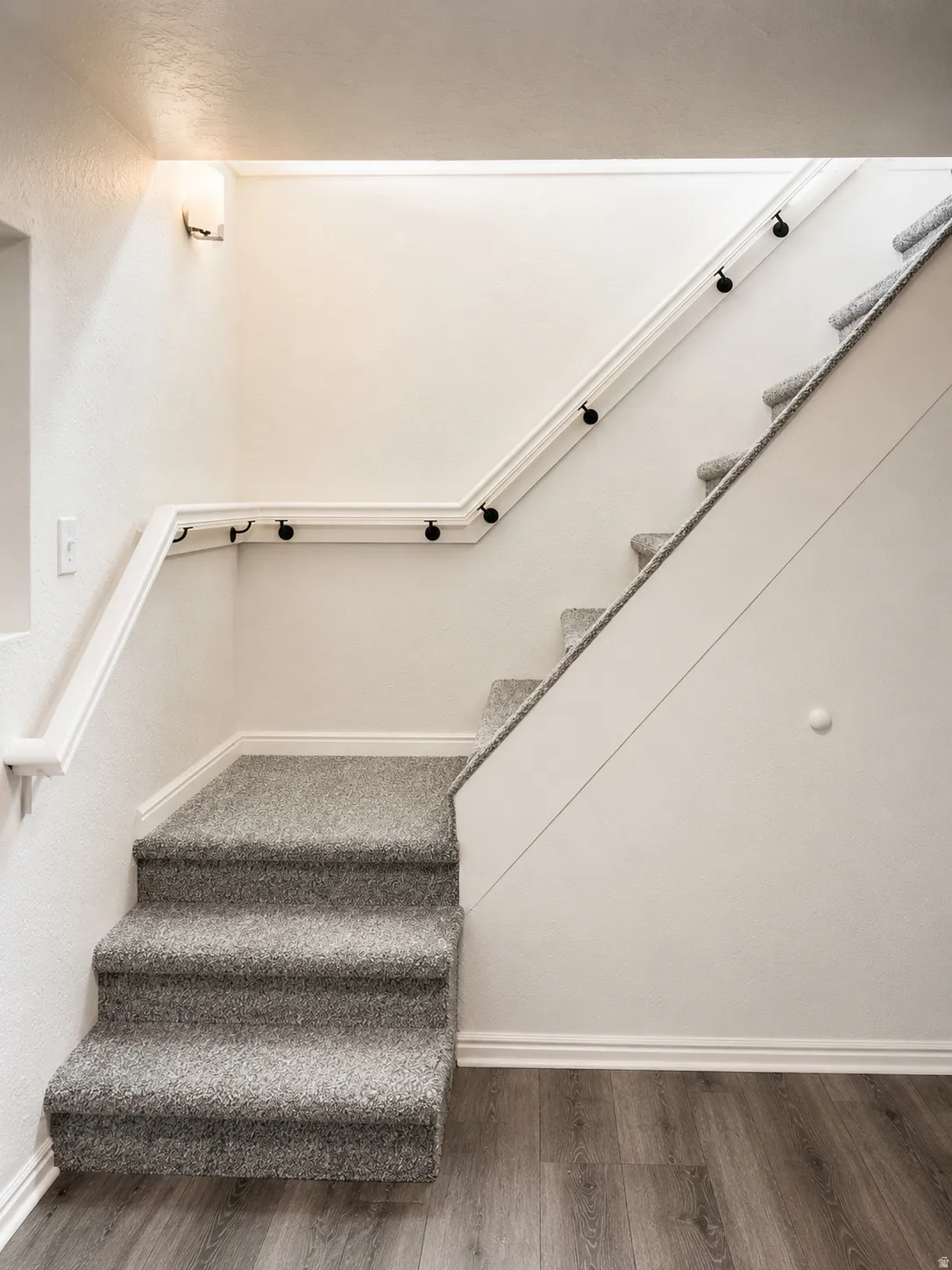 Carpeted staircase with white trim, wall-mounted sconce lighting, and wood-finish flooring at the base