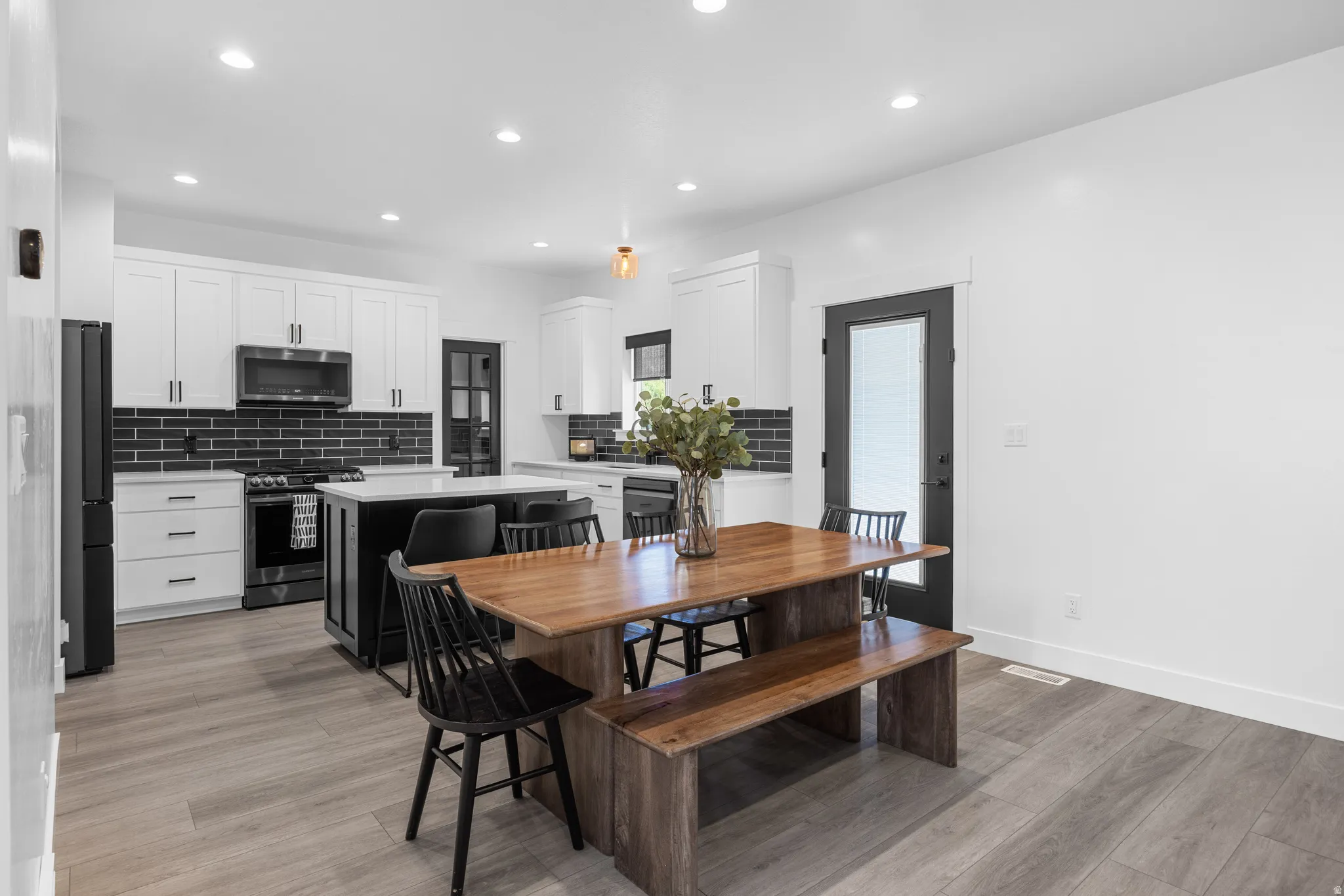 Open-concept kitchen and dining area featuring light wood-finish flooring, a kitchen island with white countertop, white cabinetry with black hardware, a black subway tile backsplash, and a black-framed door with frosted glass