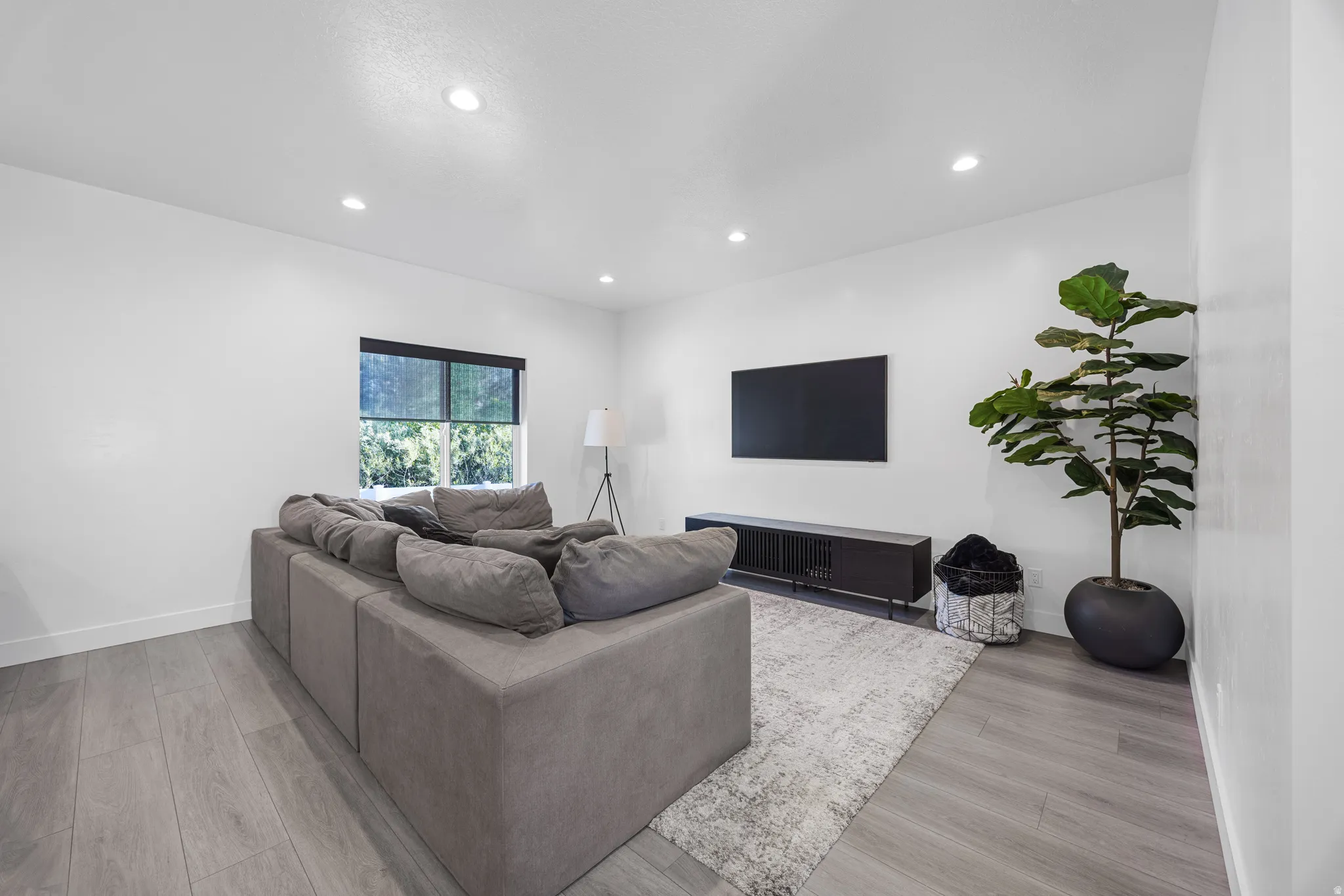 Recessed ceiling lighting illuminates a spacious room featuring light wood-finish flooring, white wall finishes, and a dark-framed window