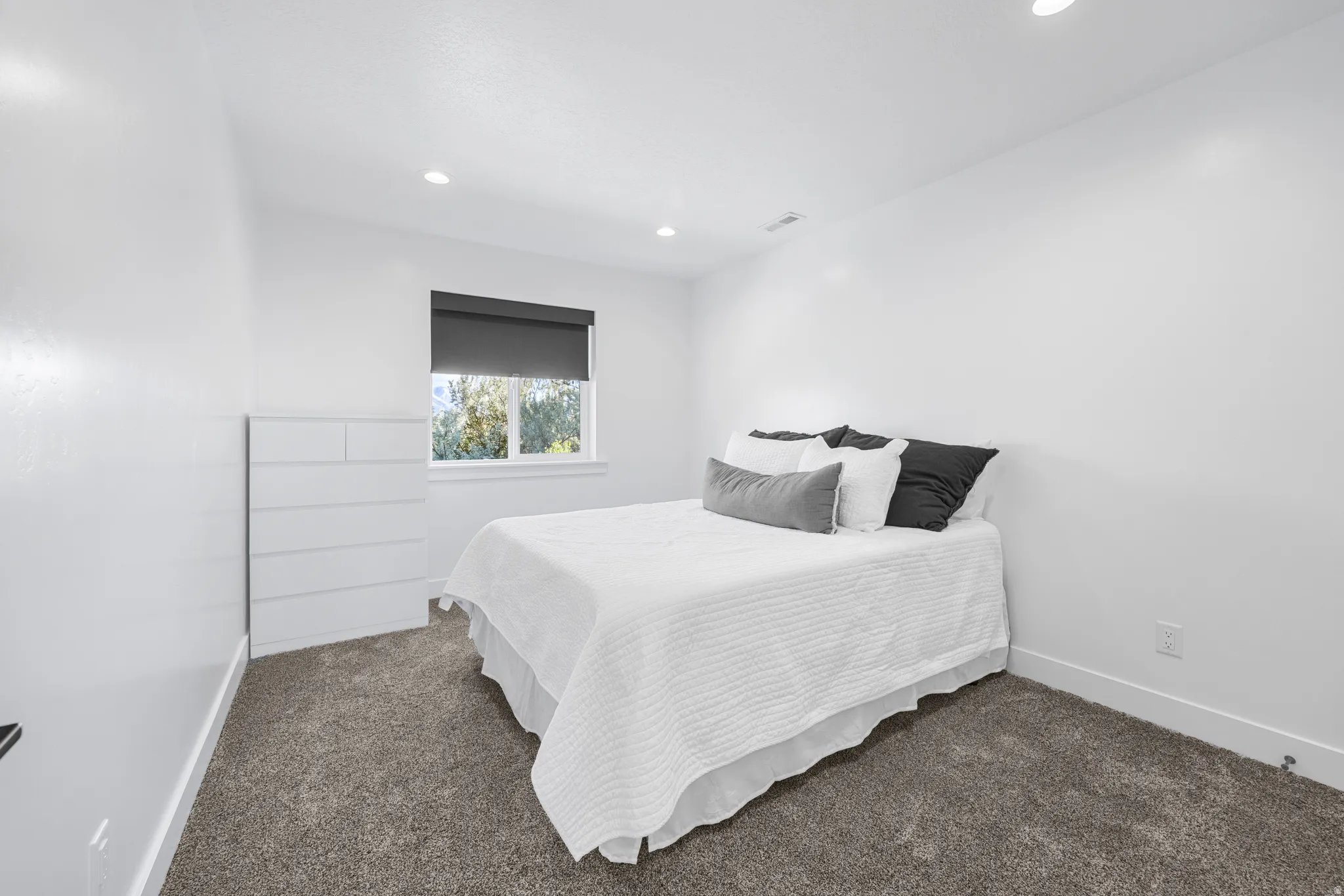 Bedroom featuring plush gray carpeting, recessed lighting, white walls, and a window with a dark roller shade