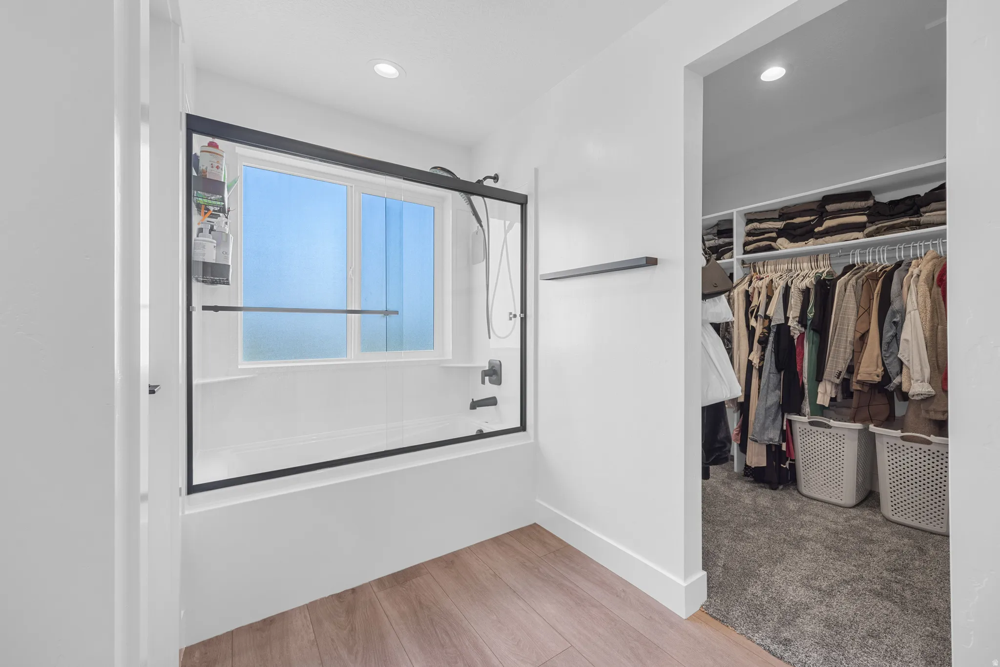 Bathroom featuring wood-finish flooring, a white-tiled tub with a black-framed glass enclosure, and a window for natural light