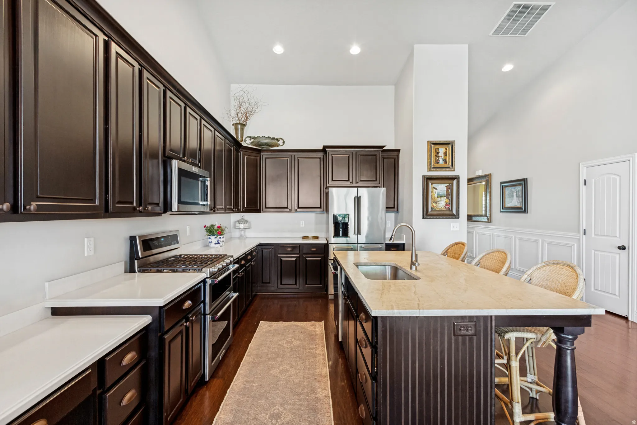 Kitchen featuring dark wood cabinetry, light-toned countertops, stainless steel appliances, a center island with sink, and hardwood flooring