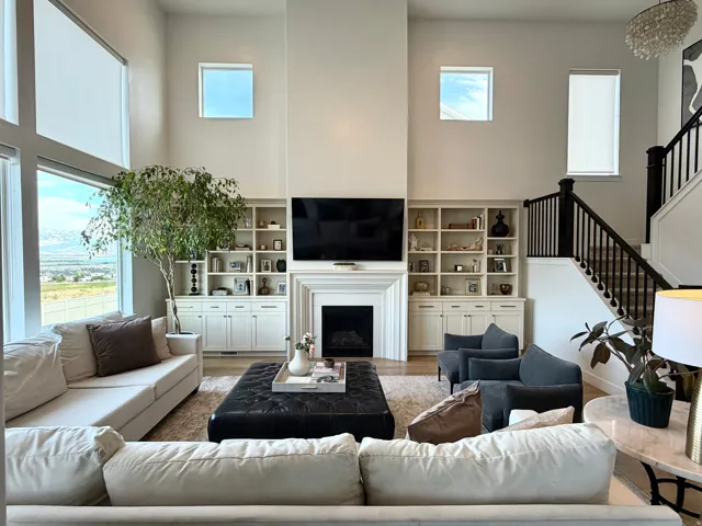 Two-story living area featuring a fireplace with mantle, built-in shelving and cabinetry, substantial window banks, and a staircase with dark wood newel posts and railings