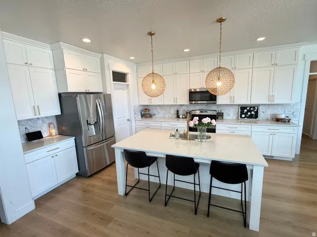 Kitchen featuring white shaker cabinetry, stainless steel appliances, a white island with seating, a marble-look tile backsplash, and wood-finish flooring