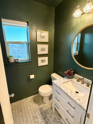 Herringbone pattern tile flooring in a bathroom featuring a white vanity with a light-toned countertop, a gold-framed mirror, and an updated light fixture