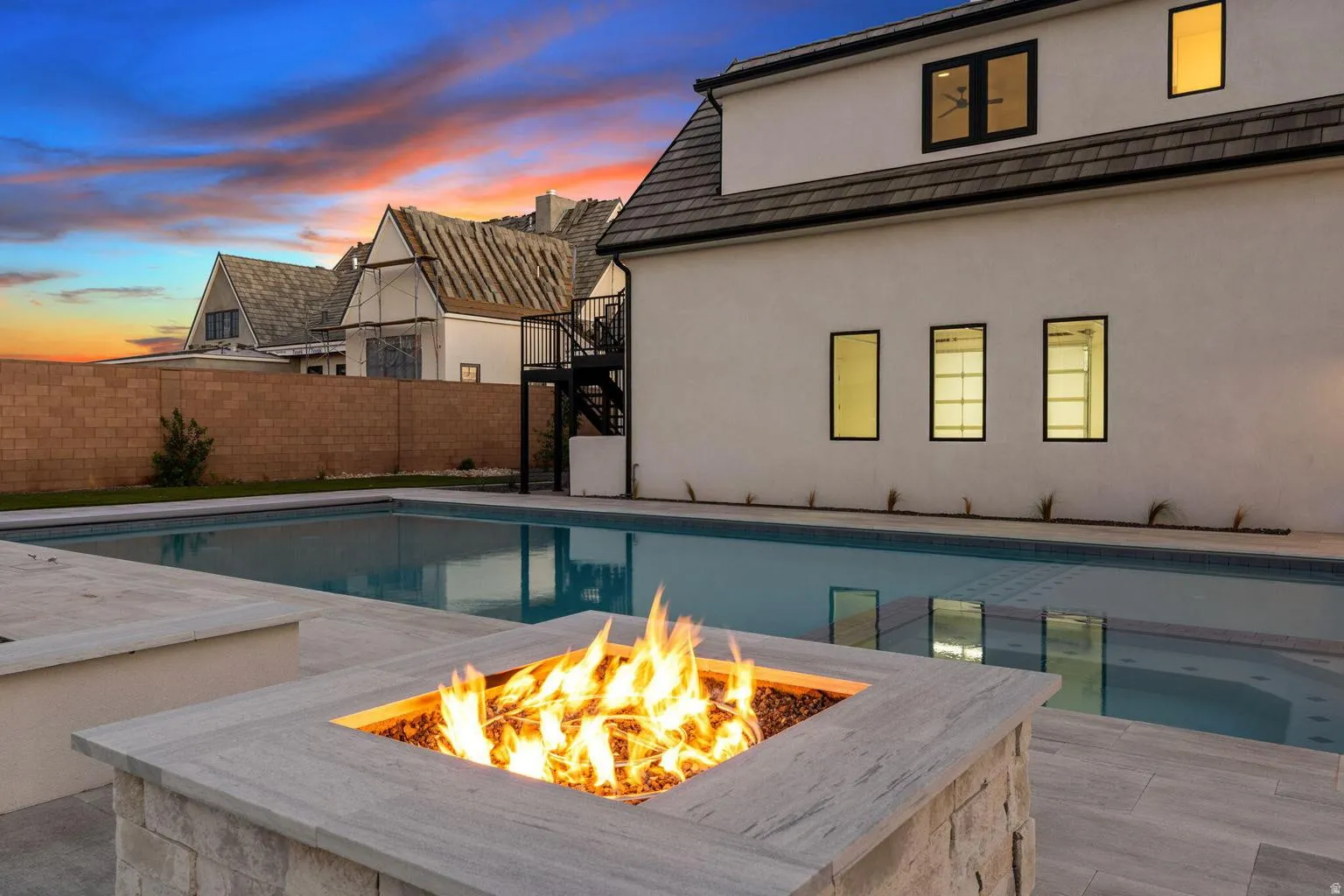 Fire pit with a stone veneer base and a square concrete cap, positioned alongside a rectangular swimming pool with a Baja shelf