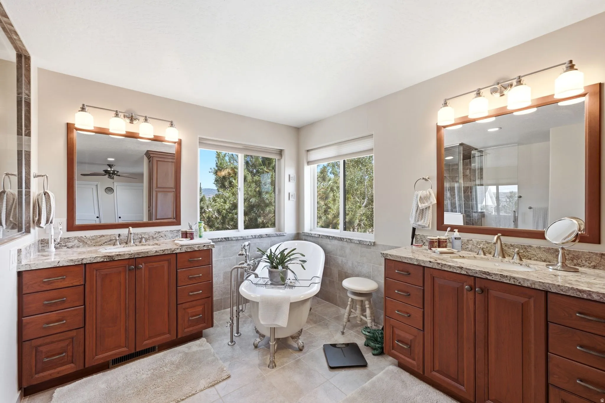 Freestanding clawfoot tub with dual windows, flanked by two wood-finish vanities with light stone countertops, integrated sinks, and framed mirrors