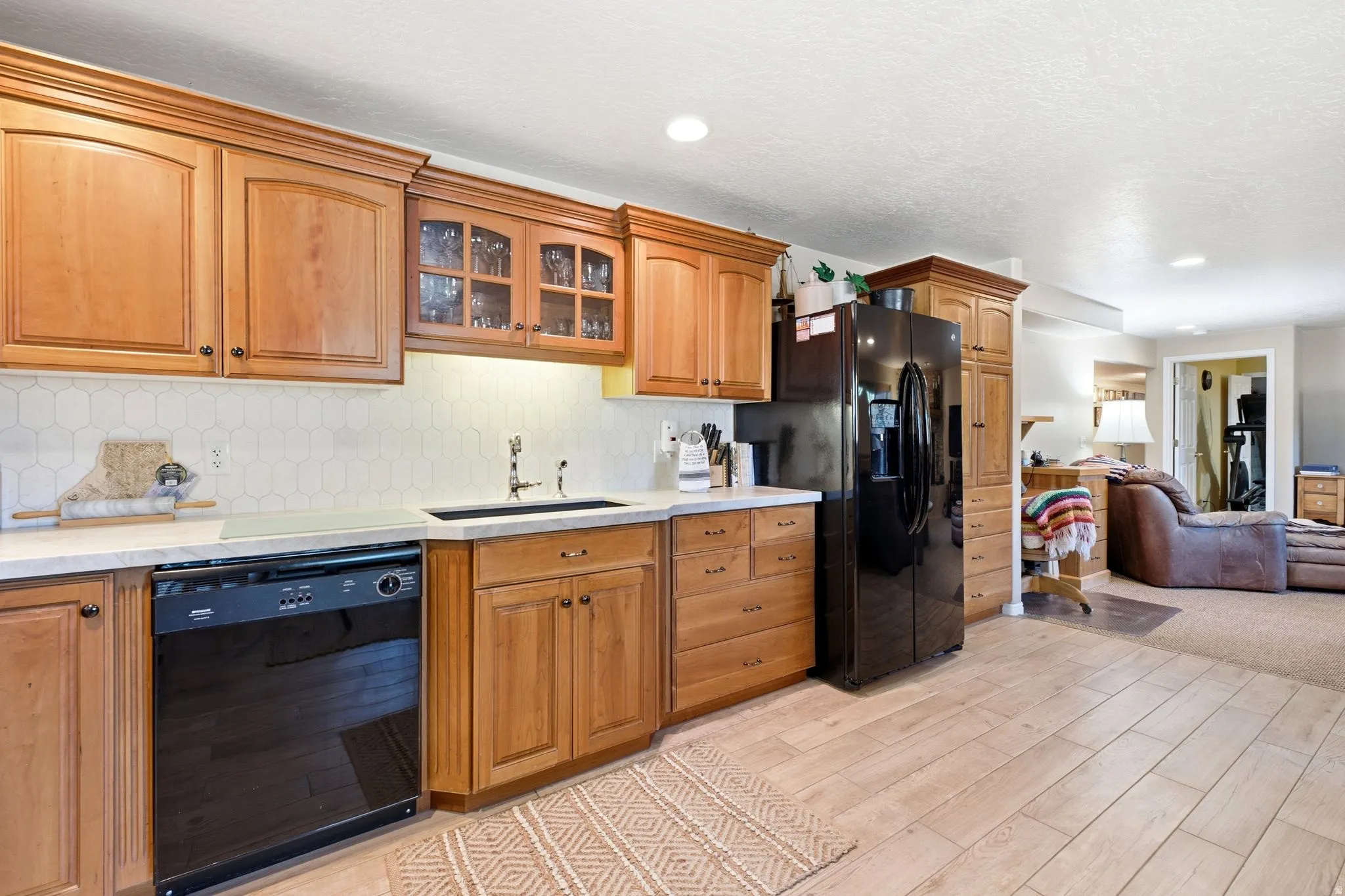 Lower level Kitchen area featuring wood cabinetry, a white hexagonal tile backsplash, light-toned countertops, and a black appliance suite