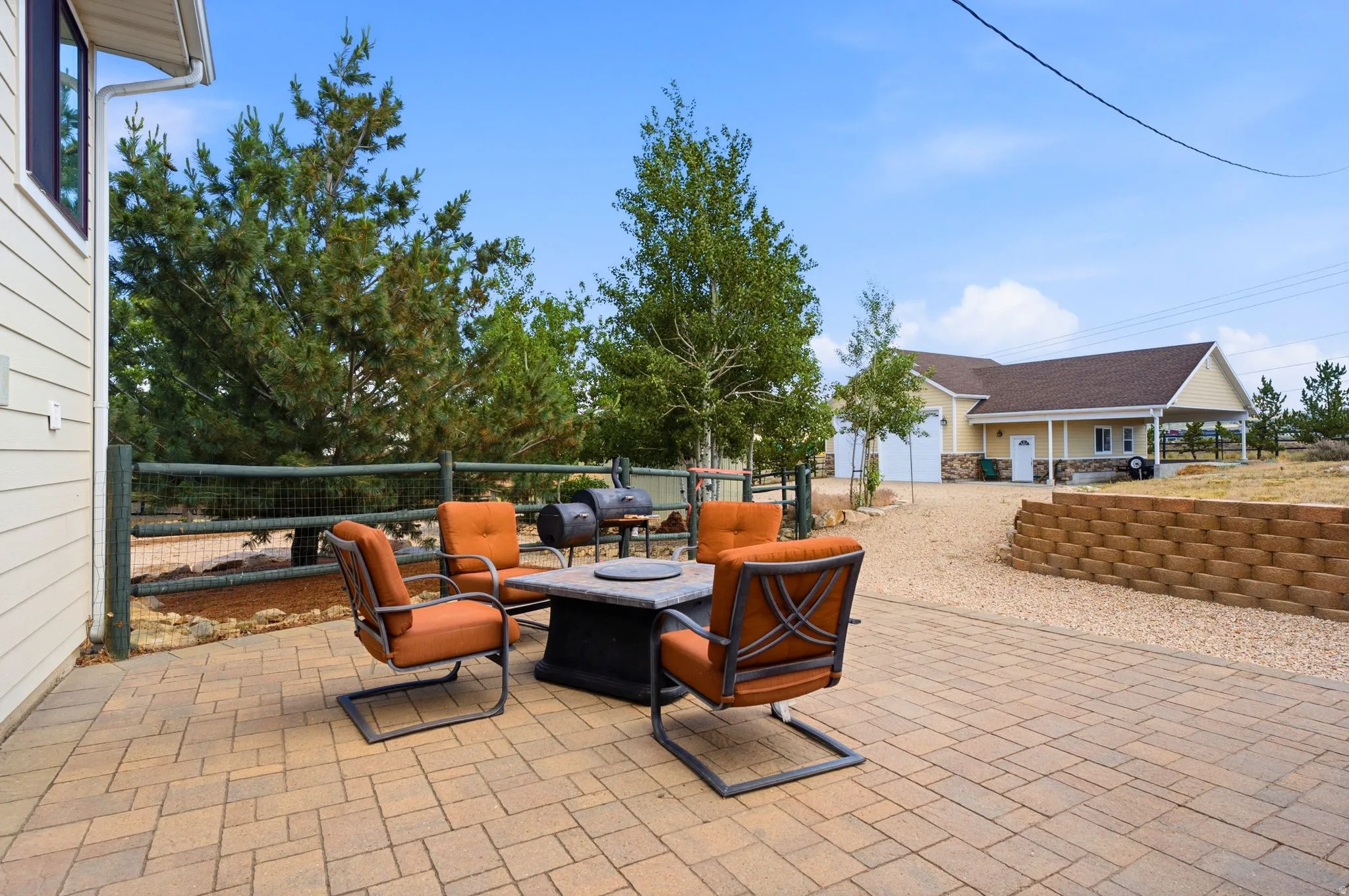 Spacious outdoor patio area featuring block pavers, a stone retaining wall, mature trees, and a gravel driveway leading to a detached garage structure with a covered carport