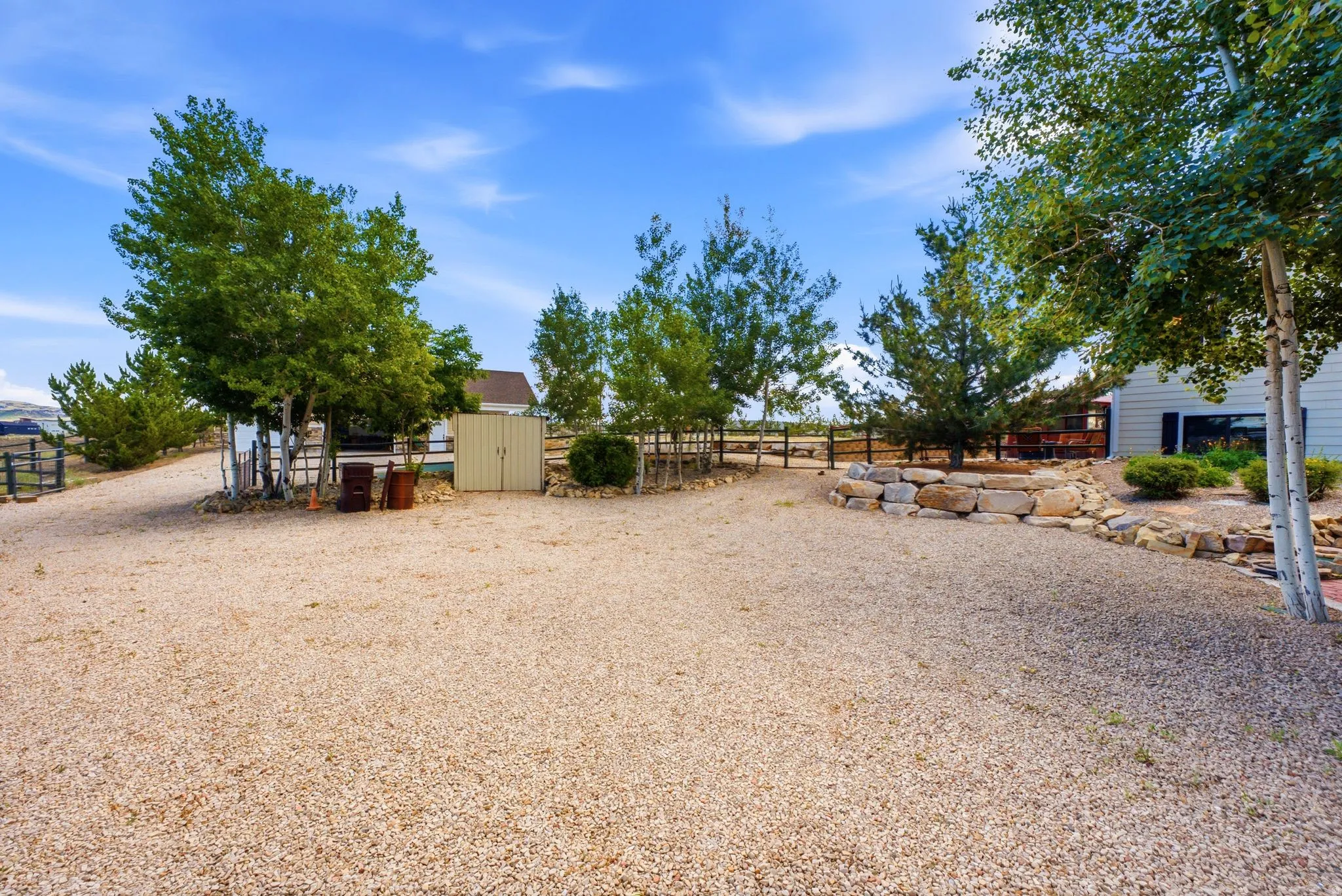 Expansive gravel driveway featuring a rock retaining wall, mature trees, a utility shed, and partial fencing