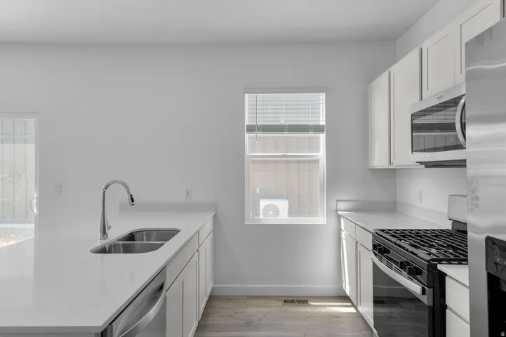 Contemporary kitchen featuring white cabinetry, light-toned countertops, a stainless steel double basin sink with a gooseneck faucet, a stainless steel dishwasher, and a stainless steel gas range with an overhead microwave