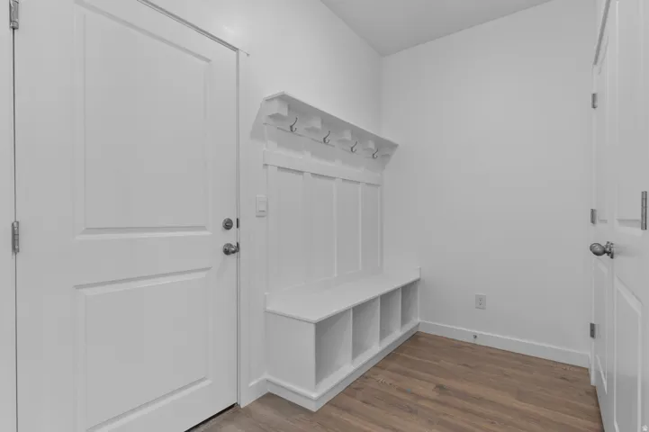Entryway featuring a built-in white bench with integrated storage cubbies, white wall paneling with shelving and metal coat hooks, and wood-finish flooring
