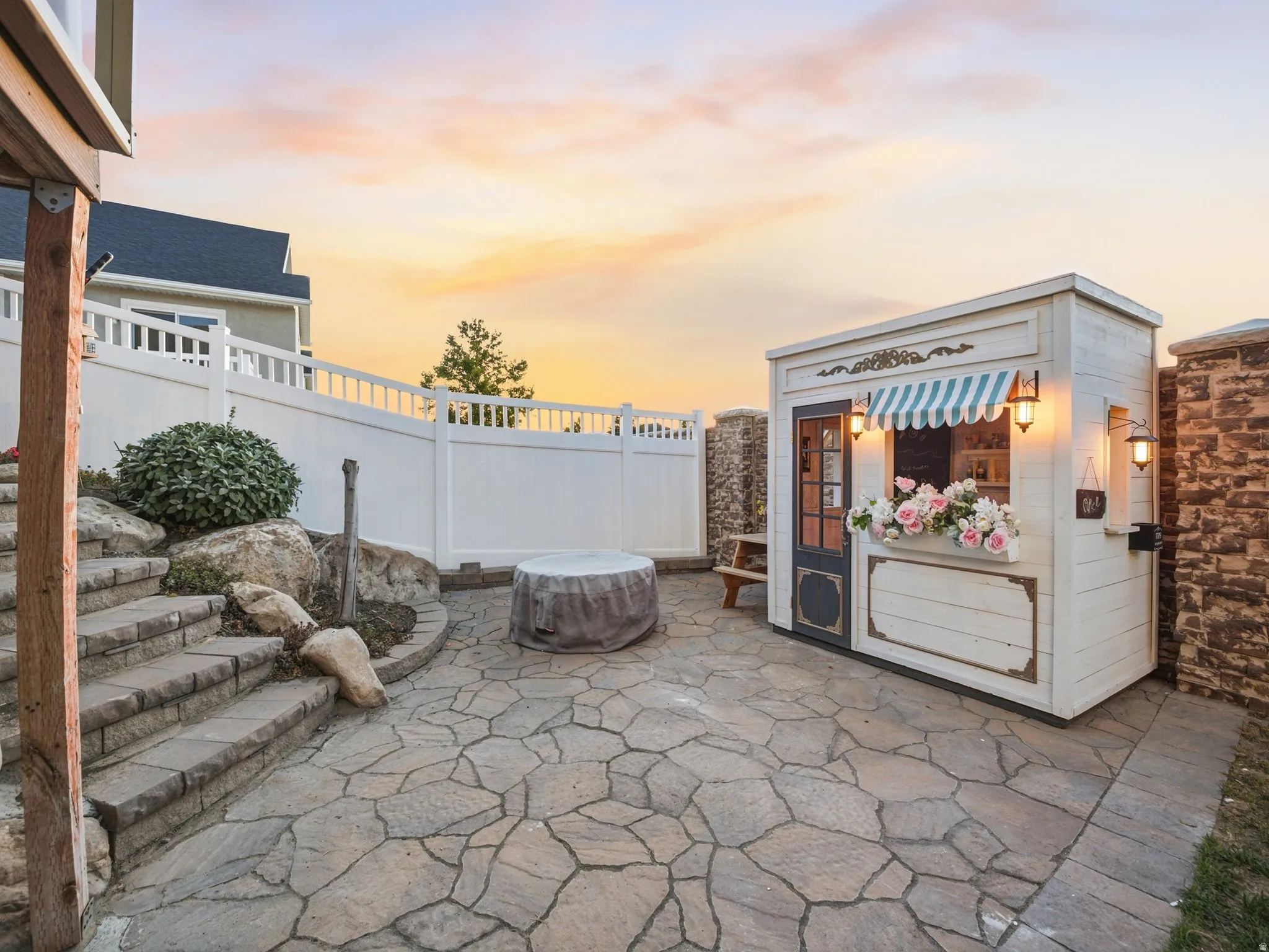 Stone patio featuring a built-in playhouse with an awning and sconce lighting, bordered by a white privacy fence and retaining walls