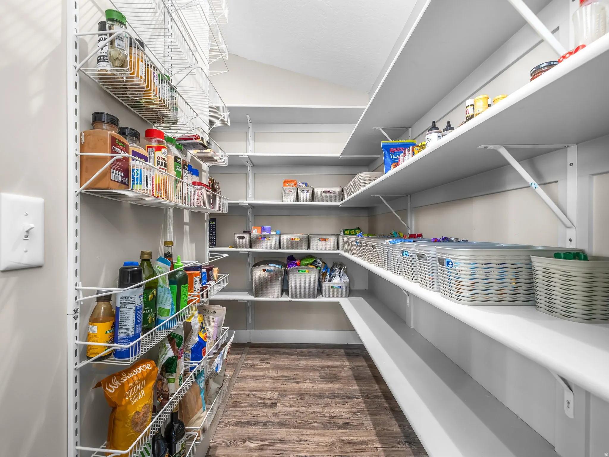 Spacious pantry featuring extensive white shelving and wire racks, wood-finish flooring, and light gray wall paint