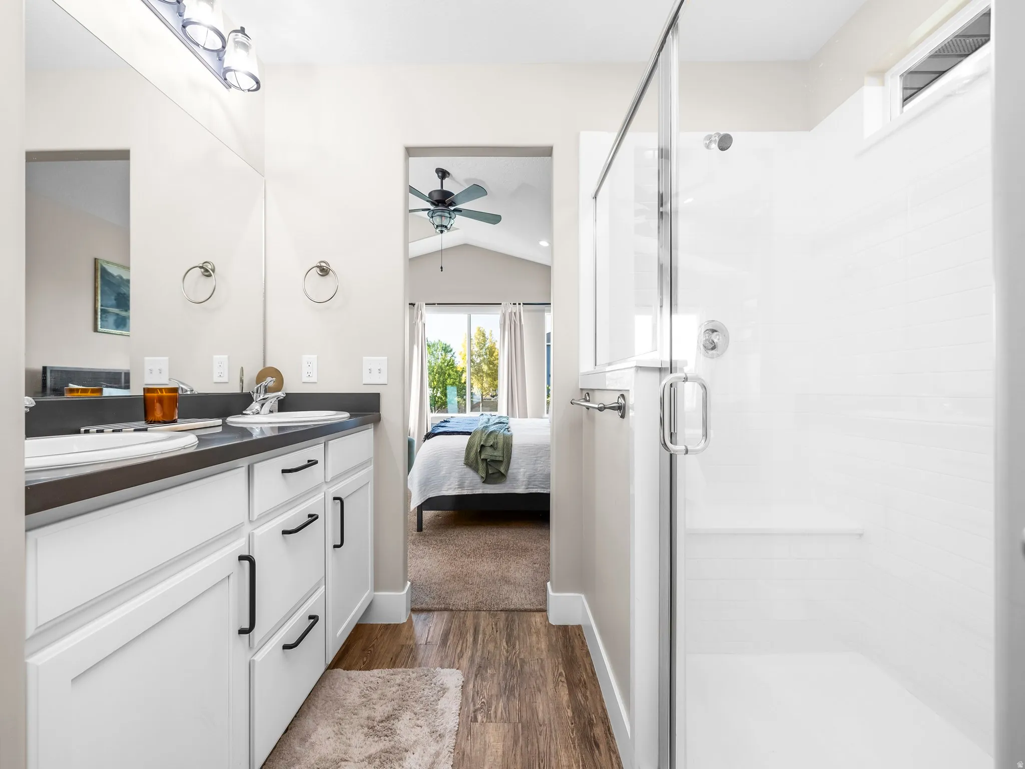 Dual vanity with dark countertops and white cabinetry, a glass-enclosed shower with white tiled walls, and wood-finish flooring