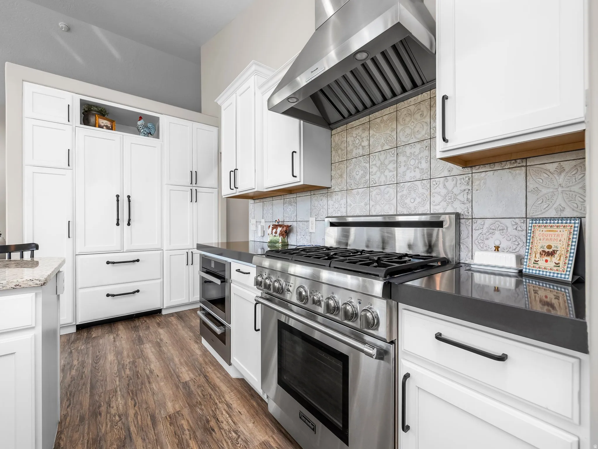 Kitchen featuring white shaker cabinetry, stainless steel range with matching range hood, dark countertops, patterned tile backsplash, and wood-finish flooring