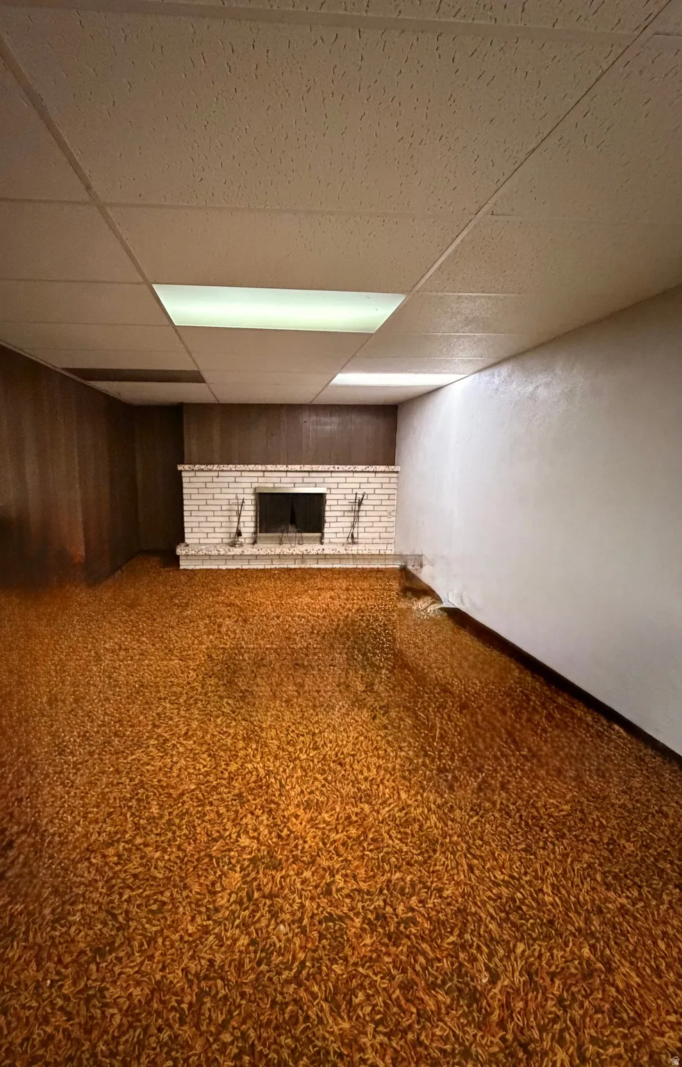 Spacious room featuring a brick fireplace with hearth, wood paneling, acoustic tile ceiling, and integrated fluorescent lighting