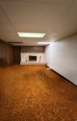 Spacious room featuring a brick fireplace with hearth, wood paneling, acoustic tile ceiling, and integrated fluorescent lighting
