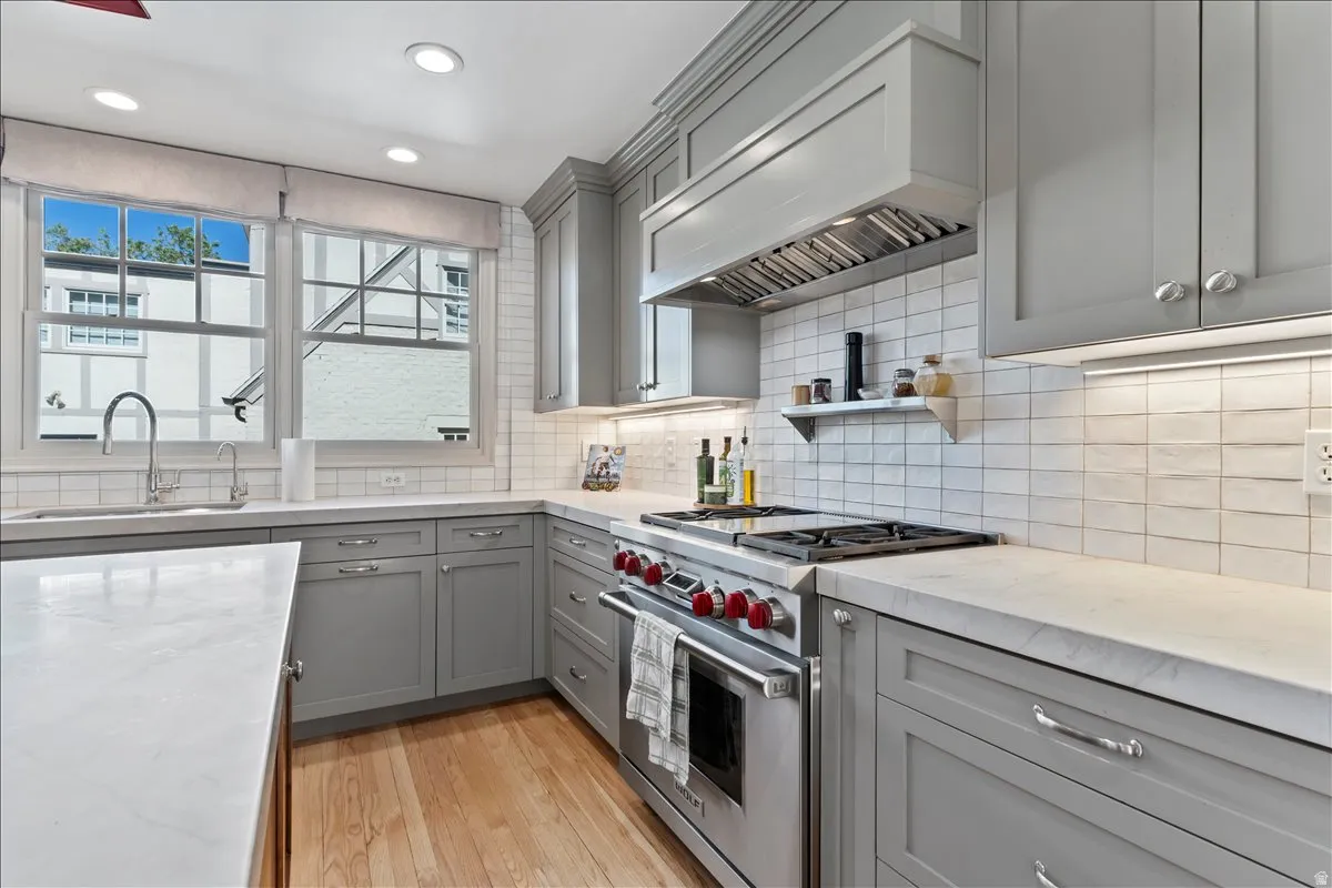 Kitchen featuring light gray shaker cabinetry, white tile backsplash, light stone countertops, stainless steel gas range, and wood-finish flooring