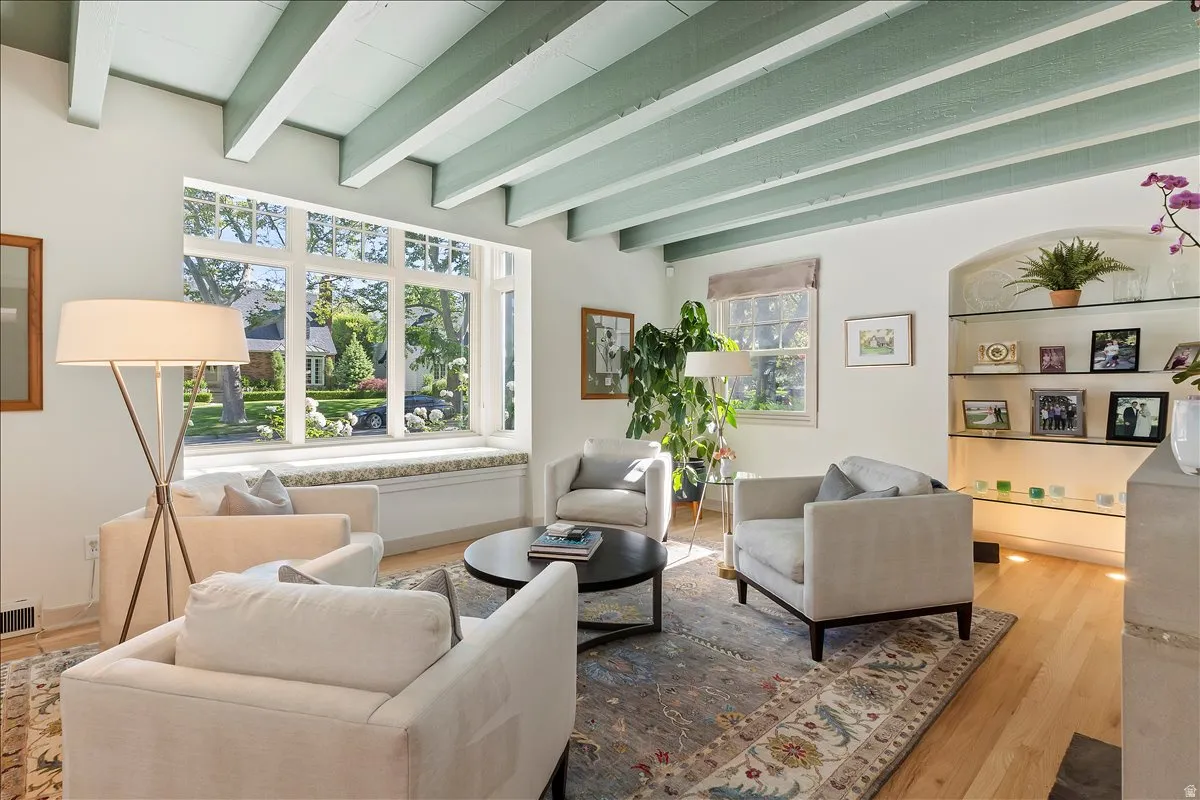 Living area featuring a large picture window with a built-in window seat, exposed ceiling beams, hardwood flooring, and an arched alcove with glass shelving and integrated lighting