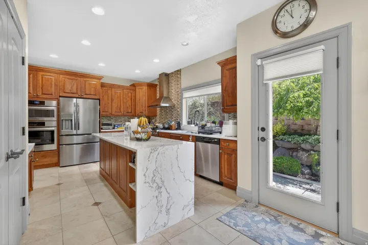 Kitchen featuring rich wood cabinetry, stainless steel appliances, a waterfall island with stone countertop, tile flooring, and a glass-paneled door