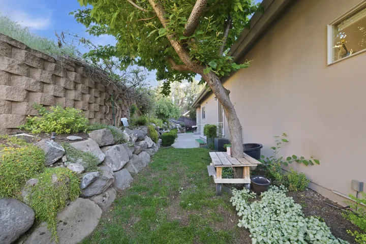 Lush side yard featuring a decorative retaining wall, natural rock landscaping, mature tree, manicured lawn, and stucco exterior