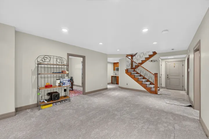 Spacious carpeted area with recessed lighting, an open wood staircase with wrought iron balusters, a built-in wet bar with wood cabinetry and a stainless steel sink, and a solid panel door