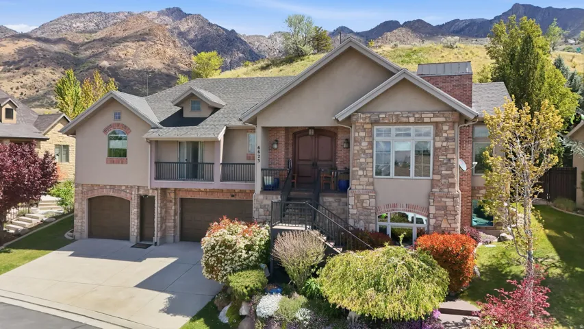 Multi-level residence featuring a stone and stucco facade, an integrated two-car garage, a front entry balcony with black railing, and mature landscaping