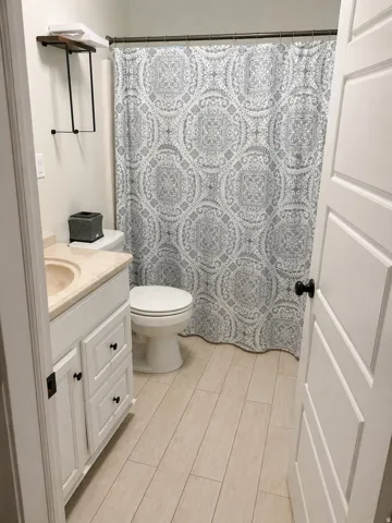 Bathroom with light wood-finish plank flooring, a white vanity with paneling and dark hardware, a light-colored countertop with an integrated sink, and a commode